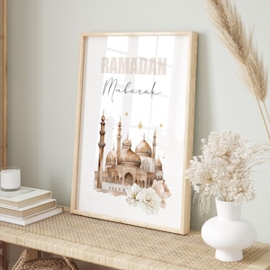 May include: A framed art print with a watercolor illustration of a mosque, featuring brown domes and minarets. The words "Ramadan Mubarak" are printed at the top. The artwork is decorated with white and pink flowers and gold stars.
