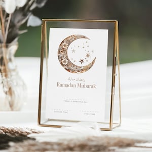 Puede incluir: Una tarjeta con marco dorado con un diseño de luna creciente y estrellas, con las palabras "Ramadan Mubarak" en inglés y árabe. La tarjeta está sobre una superficie blanca, con un fondo borroso.