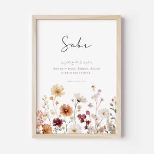 May include: A framed print featuring the word "Sabr" in elegant script above Arabic text and English translation: "And be patient. Indeed, Allah is with the patient." The bottom of the print is adorned with a floral arrangement in warm tones.