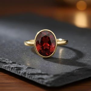 May include: A gold ring with a large, oval-shaped, deep red gemstone. The gemstone is set in a bezel setting, and the band is polished gold. The ring is shown on a dark, textured surface.