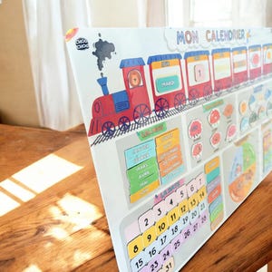 Puede incluir: Un calendario infantil con un diseño de tren rojo. El calendario muestra los días de la semana, los meses y las fechas. El texto "MON CALENDRIER" está en la parte superior. Medidas en centímetros.