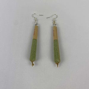 May include: A pair of novelty earrings shaped like rolled cigarettes. Each earring features a green, cylindrical body with a tan paper tip and a silver fish hook ear wire. The earrings are displayed against a white background.