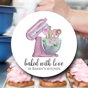 May include: A round, white graphic with a pink stand mixer, baking tools, and floral accents. The text "baked with love" and "in Sarah's kitchen" are written in black script. Cupcakes with pink frosting are in the foreground.