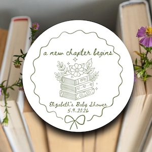 May include: A white circular sticker with a scalloped edge features the text "a new chapter begins" and "Elizabeth's Baby Shower 5.9.2026". The design includes a stack of books, flowers, leaves, and a bow.