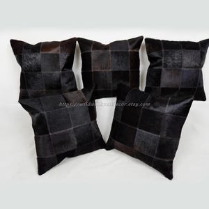 May include: Four black and gray patchwork leather throw pillows. The pillows are arranged in a row on a white surface. The pillows are made of cowhide and have a unique, textured look.