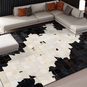 May include: A black and white cowhide rug with a patchwork design. The rug is laid out on a light wood floor with a white sectional sofa in the background.