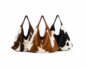 May include: Three cowhide tote bags in brown, tan, and black with white patches. Each bag has a triangular shape and a single, long strap. The bags are displayed against a white background.