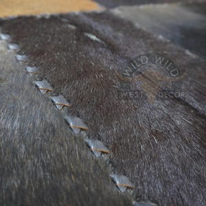 Op de afbeelding: Close-up van een grijs en bruin koeienhuidkleed met een gestikt ontwerp. Het kleed heeft een subtiele glans en een zacht, getextureerd oppervlak. De tekst "WILD WEST DECOR" is zichtbaar in de rechterbenedenhoek.