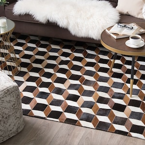 Pode incluir: Um tapete com padrão geométrico com um design de cubo 3D em preto, castanho e branco. O tapete é feito de um material macio e é colocado num chão de madeira. O tapete é um elemento decorativo para uma sala de estar.