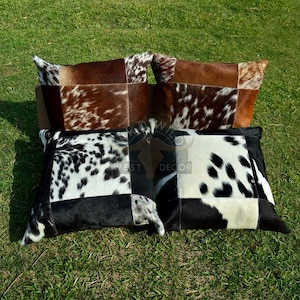 Könnte beinhalten: Vier dekorative Kuhfellkissen in Braun-, Schwarz- und Weißtönen sind auf grünem Gras angeordnet. Die Kissen haben ein Patchwork-Design, wobei jedes Kissen ein einzigartiges Muster aus Flecken und Flicken aufweist.
