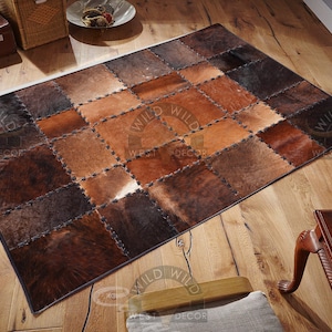 以下が含まれることがあります： 茶色、ベージュ、黒の牛革で作られたパッチワークの敷物。敷物は、さまざまなサイズと色の正方形で構成されています。敷物は木製の床の上にあります。