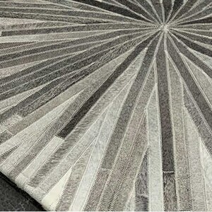 Pu&ograve; includere: Un tappeto in pelle di mucca grigio e bianco con un design circolare. Il tappeto &egrave; realizzato con strisce di pelle cucite insieme per creare un motivo a raggiera.