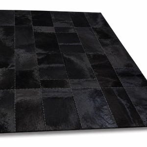 Può includere: Un tappeto rettangolare composto da diversi pannelli di pelle bovina nera cuciti insieme. Il tappeto presenta una consistenza naturale e liscia con varie sfumature di nero profondo sui singoli pezzi di pelle uniti da cuciture visibili.