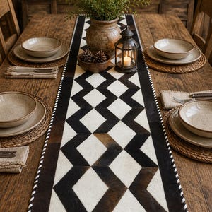 May include: A rectangular table runner featuring a geometric pattern of black, brown, and white cowhide patches. The runner has a stitched white and black border and is centered on a wooden table with place settings including bowls and napkins.