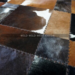 以下が含まれることがあります： 茶色、黒、白の牛革を使用したパッチワークラグ。各パーツは目に見える縫い目で縫い合わされています。「WILD WILD WEST DECOR」の文字がラグに表示されています。