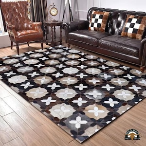 May include: A rectangular area rug with a geometric pattern in shades of brown, black, white, and tan. The rug is placed on a hardwood floor, with a leather sofa and armchair in the background. The rug has a repeating floral-like design.