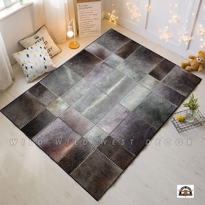 May include: A patchwork cowhide rug with a grey, brown, and white colour scheme. The rug is made of individual squares of cowhide that are sewn together. The rug is laid out on a light wood floor.