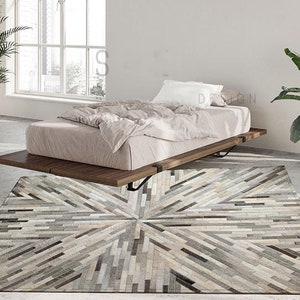 May include: A gray and white cowhide rug with a geometric pattern. The rug is laid out on a light gray floor in front of a bed with white bedding.