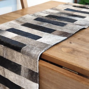 May include: A black and white patchwork table runner made of leather. The runner has a rectangular shape and is made of different sized pieces of leather sewn together.