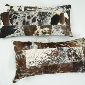 May include: Two rectangular cowhide pillows. The pillows are a patchwork of brown, black, and white fur. The pillows are on a white surface. The pillows are a decorative item for a home.