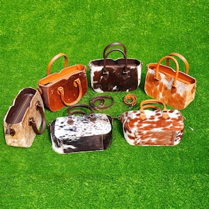 Könnte beinhalten: Eine Auswahl an Handtaschen und Accessoires aus Kuhleder in verschiedenen Farben und Mustern, darunter Braun, Weiß und Schwarz. Die Taschen haben obere Griffe und sind auf einer grünen Oberfläche angeordnet.