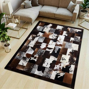 Puede incluir: Una alfombra rectangular de piel de vaca con un diseño de patchwork. La alfombra presenta cuadrados de piel de vaca marrón, negra, blanca y gris, creando un patrón geométrico. La alfombra tiene un borde marrón oscuro y está colocada sobre un suelo de madera clara.