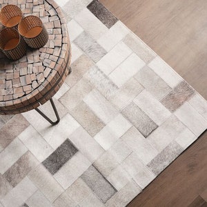 May include: A white and gray cowhide rug with a geometric pattern. A wooden table with a mosaic top sits on the rug. Three gold candle holders are on the table.