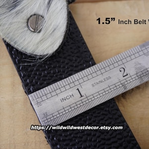 May include: Close-up of a black leather belt with a textured surface, featuring a buckle with a white and gray fur accent. The belt width is 1.5 inches. The Etsy shop URL is visible at the bottom.