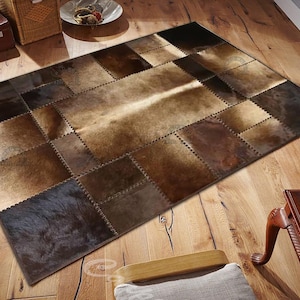 Könnte beinhalten: Ein brauner und beigefarbener Patchwork-Rinderfellteppich mit einem Used-Look. Der Teppich besteht aus verschiedenen Lederstücken, die zusammengenäht wurden, um ein einzigartiges Design zu schaffen.