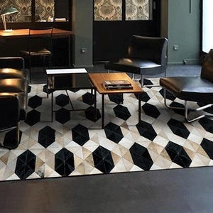 May include: A geometric patterned area rug in a modern living room setting. The rug features a repeating pattern of black, white, and tan shapes. The room includes black leather furniture and wooden tables, creating a stylish, contemporary space.