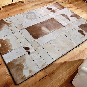 May include: Rectangular cowhide rug with a patchwork design. The rug features alternating squares of brown and white cowhide, stitched together with dark thread. The rug is placed on a hardwood floor.