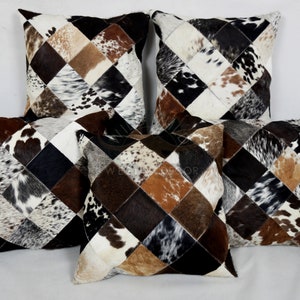 Peut inclure: Cinq coussins carrés avec un motif en losange en patchwork de cuir de vache. Les coussins sont dans des tons de brun, noir et blanc.