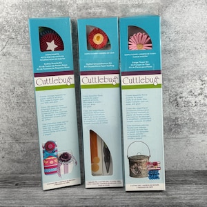May include: Three craft kits in teal boxes with floral designs. The kits are labelled "Sample Rosette," "Quilled Chrysanthemum Kit," and "Fringe Flower Kit." Each box features the "Cuttlebug" logo and instructions for paper crafts.