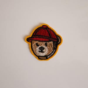 Könnte beinhalten: Gestickter Bügel-Patch mit einem Cartoon-Bären, der eine rote Baseballkappe trägt. Der Bär hat braunes Fell, eine schwarze Nase und weiße Augen.