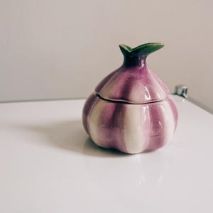 Puede incluir: Un recipiente de cerámica con forma de ajo con tapa. El recipiente es principalmente morado con rayas blancas verticales. La parte superior tiene un diseño de hoja verde. El recipiente está sobre una superficie blanca.