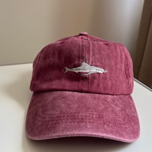 Peut inclure: Une casquette de baseball bordeaux avec un motif de poisson brodé blanc.