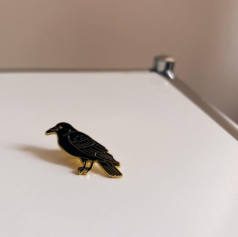 May include: A black raven enamel pin with gold-colored trim. The bird is depicted in a side profile, with detailed feather and beak features. This decorative pin is suitable for jackets, bags, or hats.