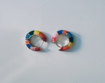 Puede incluir: Un par de pendientes de aro de colores arcoíris. Los pendientes están hechos de metal dorado y tienen un acabado de esmalte colorido. Los colores son rojo, naranja, amarillo, verde, azul y rosa.