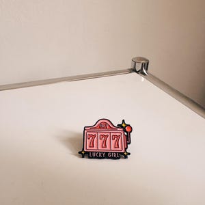 Könnte beinhalten: Eine rosa Emaille-Anstecknadel in Form eines Spielautomaten mit dem Text HEY LUCKY GIRL. Die Anstecknadel zeigt drei rote 7er in der Mitte, einen roten Hebel auf der rechten Seite und kleine gelbe Sternakzente an den Ecken, schwarz umrandet.