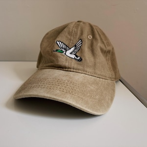 Könnte beinhalten: Beige Baseballkappe mit gebogenem Schirm. Sie hat ein gesticktes Design einer fliegenden Ente in Weiß, Schwarz und Grün. Der Hut ist aus weichem, abgenutztem Material.