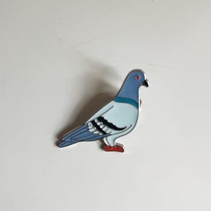 May include: A decorative enamel pin depicting a pigeon. The bird features a light blue body, a darker blue head and wings, and red feet. The pin is a colourful accessory.