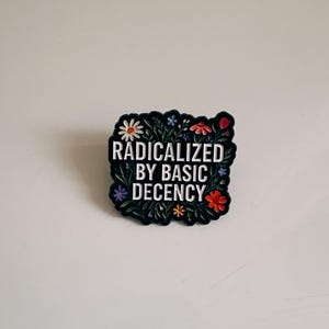 Może przedstawiać: Czarny pin z białym napisem "RADICALIZED BY BASIC DECENCY". Pin jest ozdobiony obramowaniem z zielonych liści i różnorodnych kolorowych kwiatów, w tym stokrotek, czerwonych, niebieskich i pomarańczowych kwiatów.