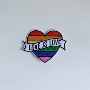 Pode incluir: Um remendo em forma de coração arco-íris com o texto "LOVE IS LOVE" em uma faixa branca.