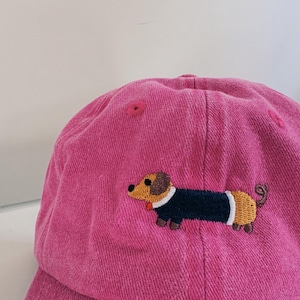 May include: A pink baseball cap featuring a dachshund dog embroidery. The embroidered dog has a brown head, a navy blue body, and a tan tail. The cap is made of a soft, textured fabric.