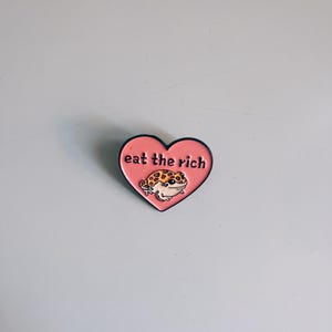 Könnte beinhalten: Ein herzförmiger Emaille-Pin in Rosa mit dem weißen Schriftzug „eat the rich“. Der Pin zeigt einen Cartoon-Gecko mit einem gepunkteten Muster. Der Pin hat eine schwarze Umrandung.