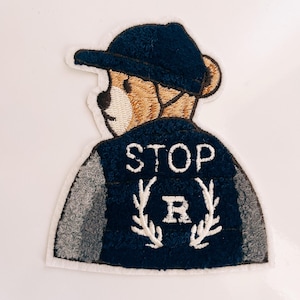 May include: Embroidered patch featuring a cartoon bear wearing a blue baseball cap and a blue jacket with a gray sleeve. The jacket has the text "STOP" and a letter "R" with a wreath design.