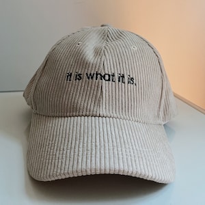 Könnte beinhalten: Beige Cord-Baseballkappe mit dem gestickten Satz "it is what it is." Die Kappe hat einen gebogenen Schirm und eine strukturierte Krone mit Belüftungsösen. Der Hut ist eine neutrale Farbe und für den Alltag geeignet.