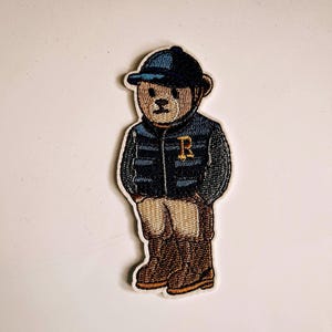 Può includere: Toppa ricamata raffigurante un orsetto con gilet trapuntato blu navy, cappellino abbinato e stivali marroni. L'orso ha la faccia color beige e la lettera "R" sul gilet.