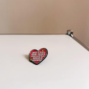 Peut inclure: Épinglette en émail en forme de cœur avec un fond rouge et une bordure noire. L'épinglette porte l'inscription "JUST A GIRL WHO NEEDS HER LITTLE SNACKS" en lettres blanches et rouges, avec des étoiles. L'épinglette est sur une surface blanche.