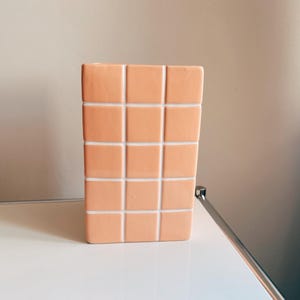 Puede incluir: Un jarrón rectangular de cerámica color melocotón con un patrón de cuadrados. Los cuadrados están delineados con líneas blancas. El jarrón está sobre una superficie blanca.